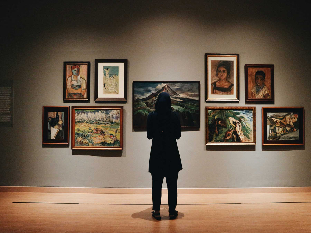 A visitor facing a salon-style hang of framed paintings on a dark wall