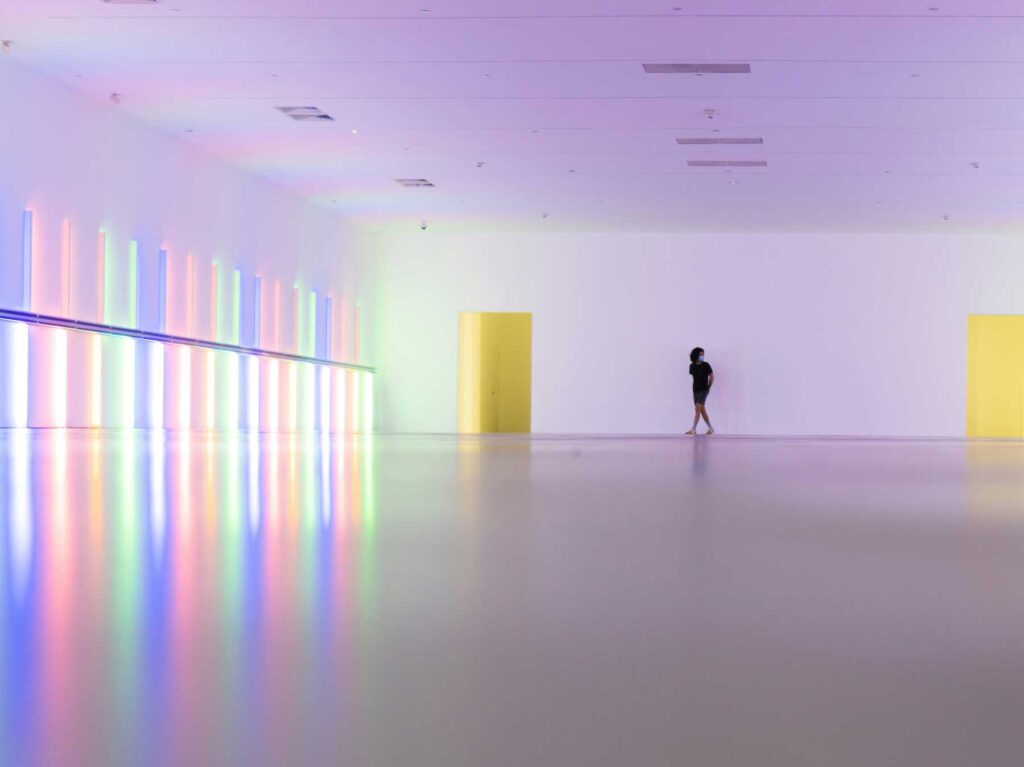 A large-scale coloured light installation filling a museum hall, with a single visitor in the distance